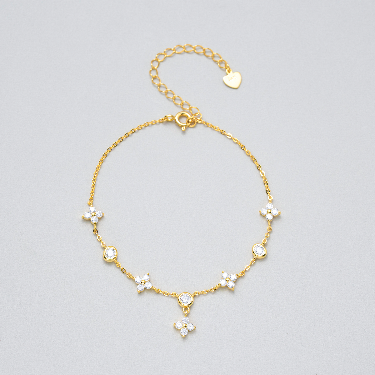 A delicate gold chain anklet featuring five small floral clusters set with clear rhinestones, a heart-shaped charm, and an adjustable clasp, displayed on a neutral light gray background.