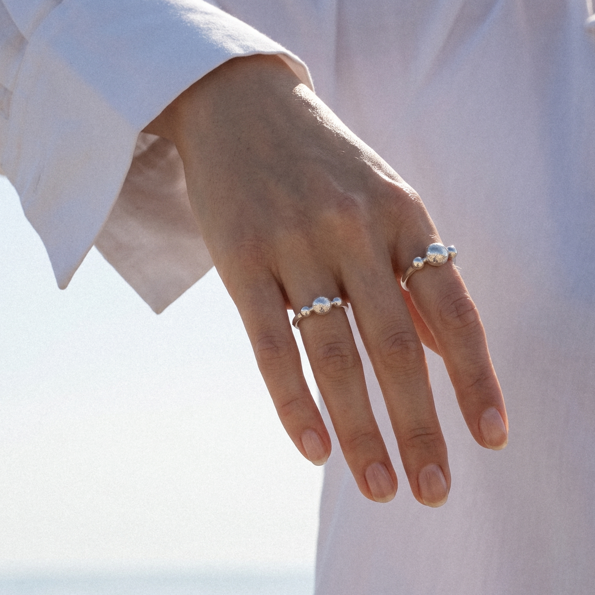 A close-up of a person's hand wearing two delicate silver rings with small white pearl-like beads, set against a soft white shirt and bright outdoor background.