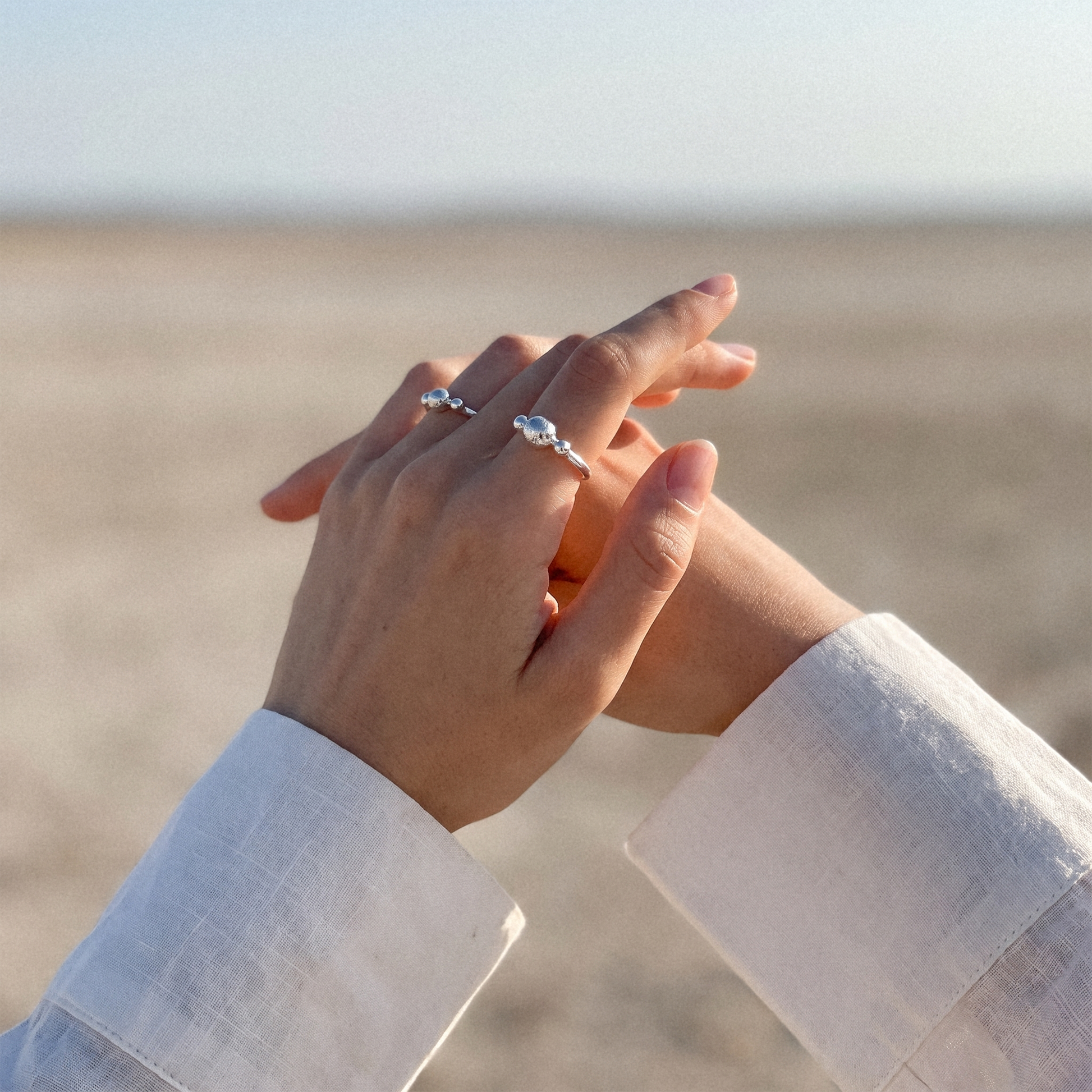 A close-up of two hands clasped together outdoors, with two silver rings featuring clear gemstones worn on the fingers. The background is a softly blurred sandy beach or desert landscape under a pale sky.