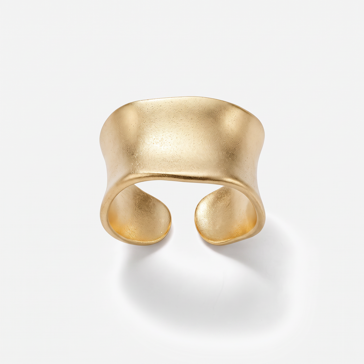 A wide, open cuff ring in matte yellow gold with a textured, organic wavy shape, photographed on a clean white background with a soft shadow.